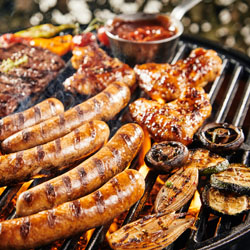 Tasty summer barbecue grilling over the hot flaming coals with sausages, steak, chicken wings marinade, garlic, onion, tomato and mushrooms